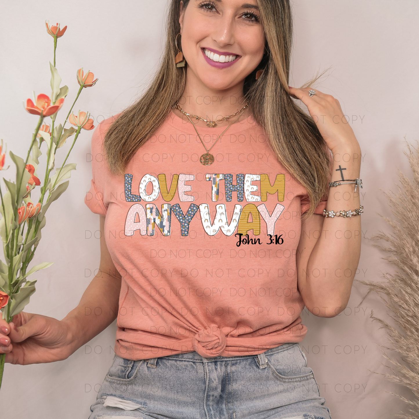 A person wearing a pink unisex t-shirt with the text 'LOVE THEM ANY WAY' printed on it, accompanied by a verse 'John 3:16'. The person is also wearing light blue jeans and accessorized with a necklace and a ring.