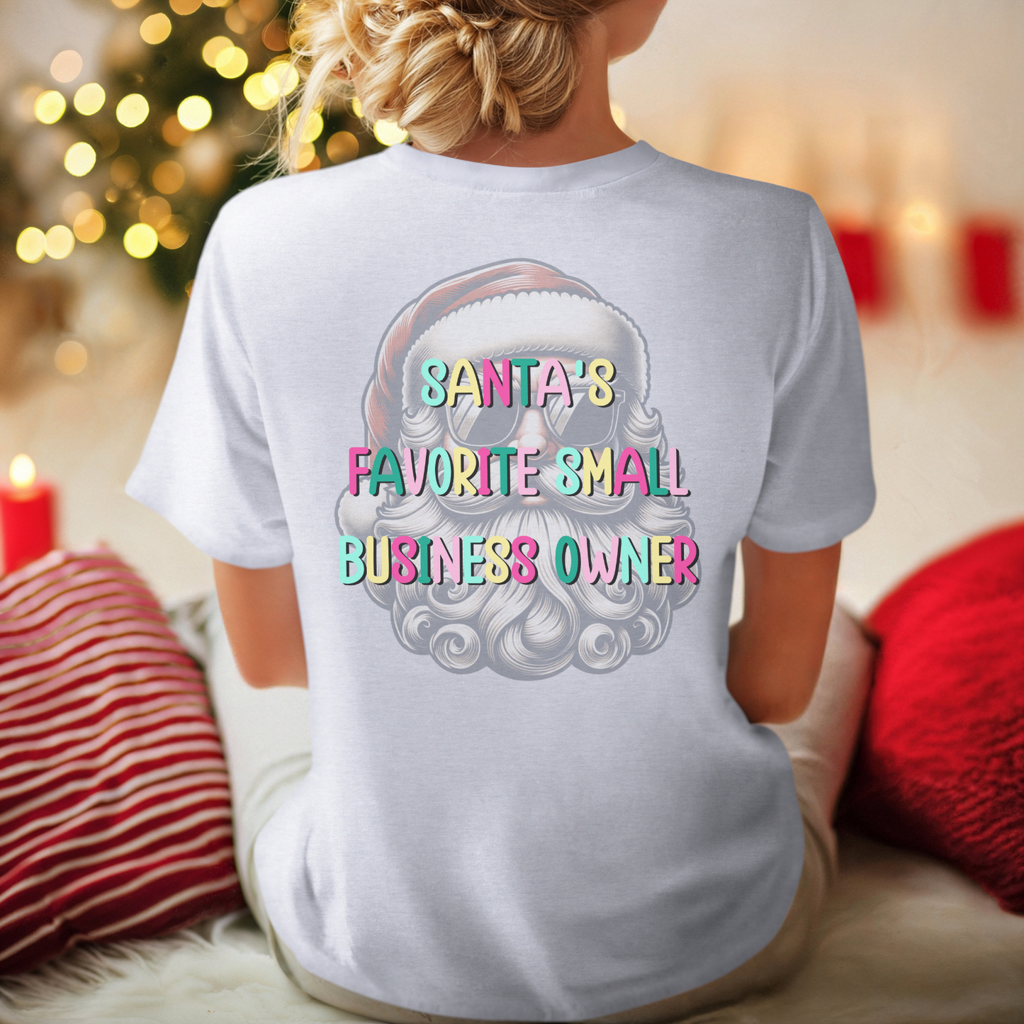 Person wearing a white t-shirt with 'Santa's Favorite Small Business Owner' text and graphic, Christmas-themed background.