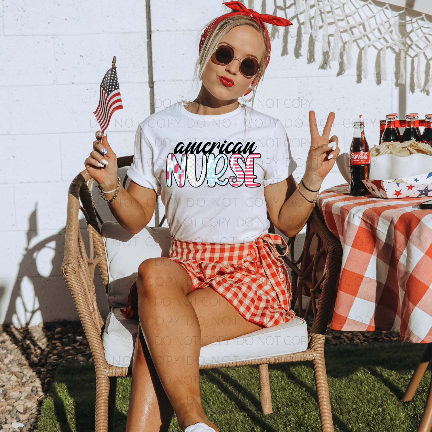 A person sitting in a chair outdoors wearing a white 'American Nurse' t-shirt with red and blue patterned shorts, holding an American flag and making a peace sign.