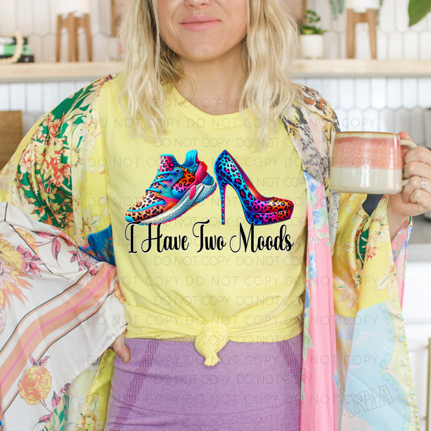 A person wearing a yellow unisex t-shirt with text and a colorful graphic of high heels on it, holding a mug.