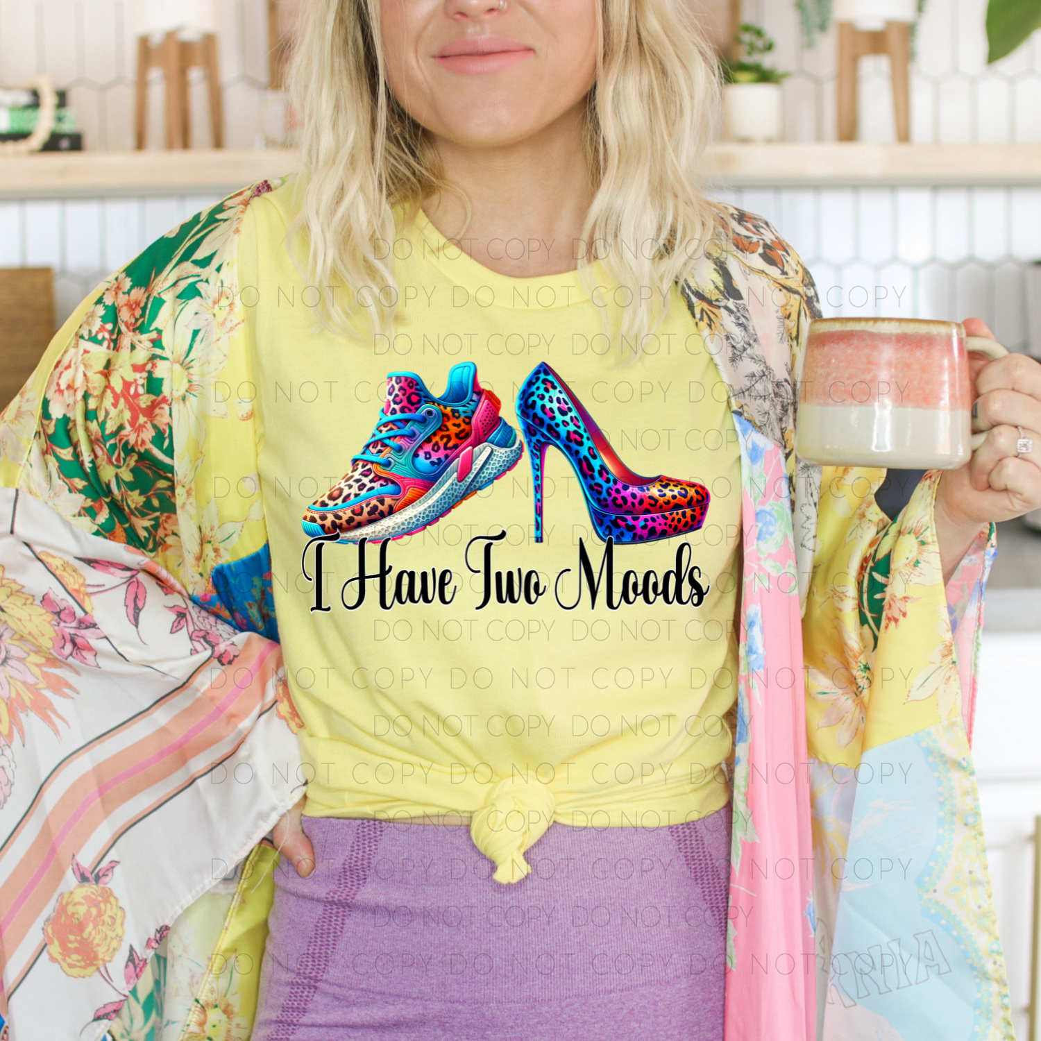 A person wearing a yellow unisex t-shirt with text and a colorful graphic of high heels on it, holding a mug.